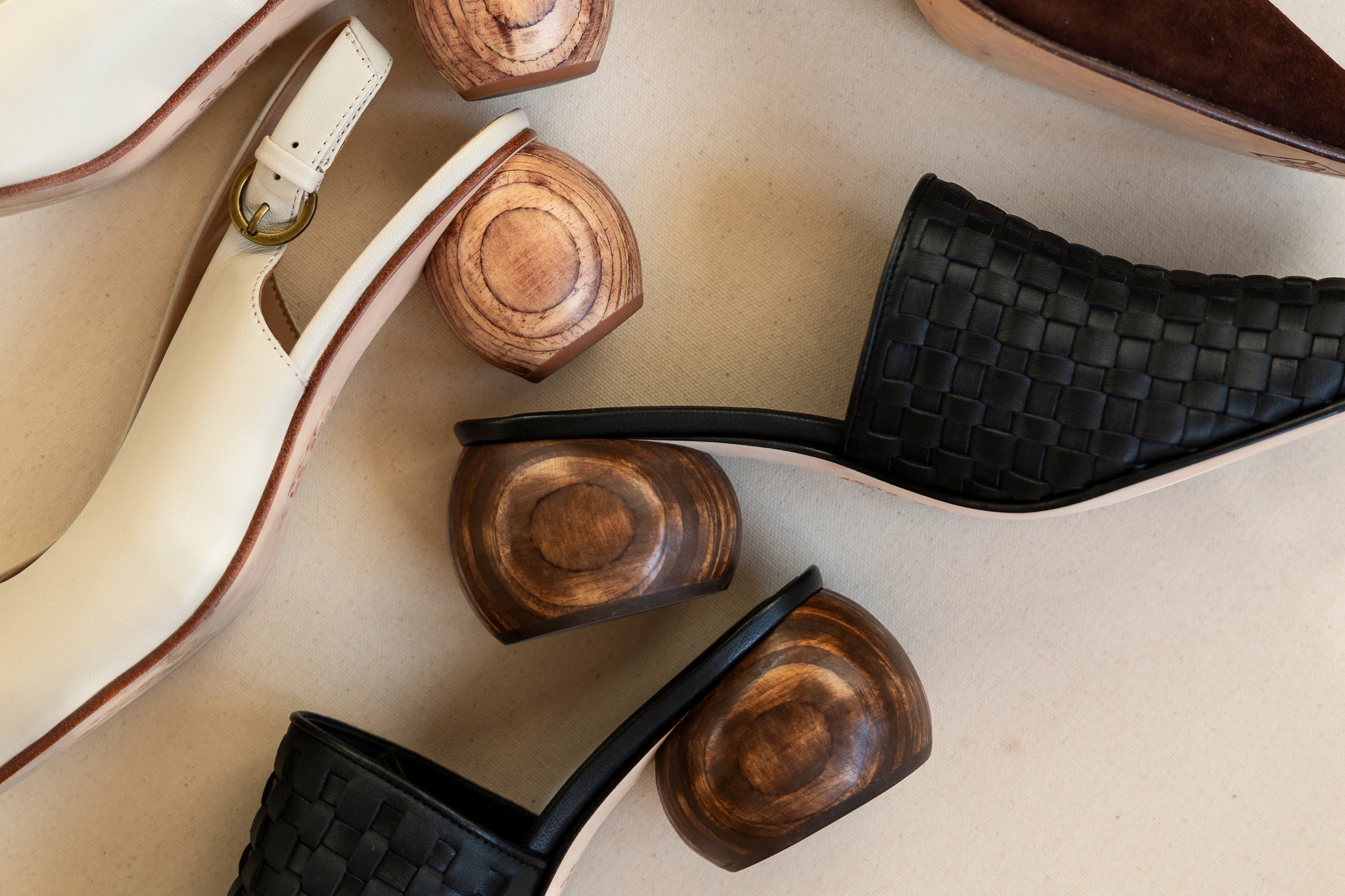 Three pairs of women's mule shoes arranged on a light beige background, each with a unique heel design: a dark brown suede pair, a cream leather pair with a white strap, and a black woven leather pair. Each pair features a chunky, sculptural wooden block heel.