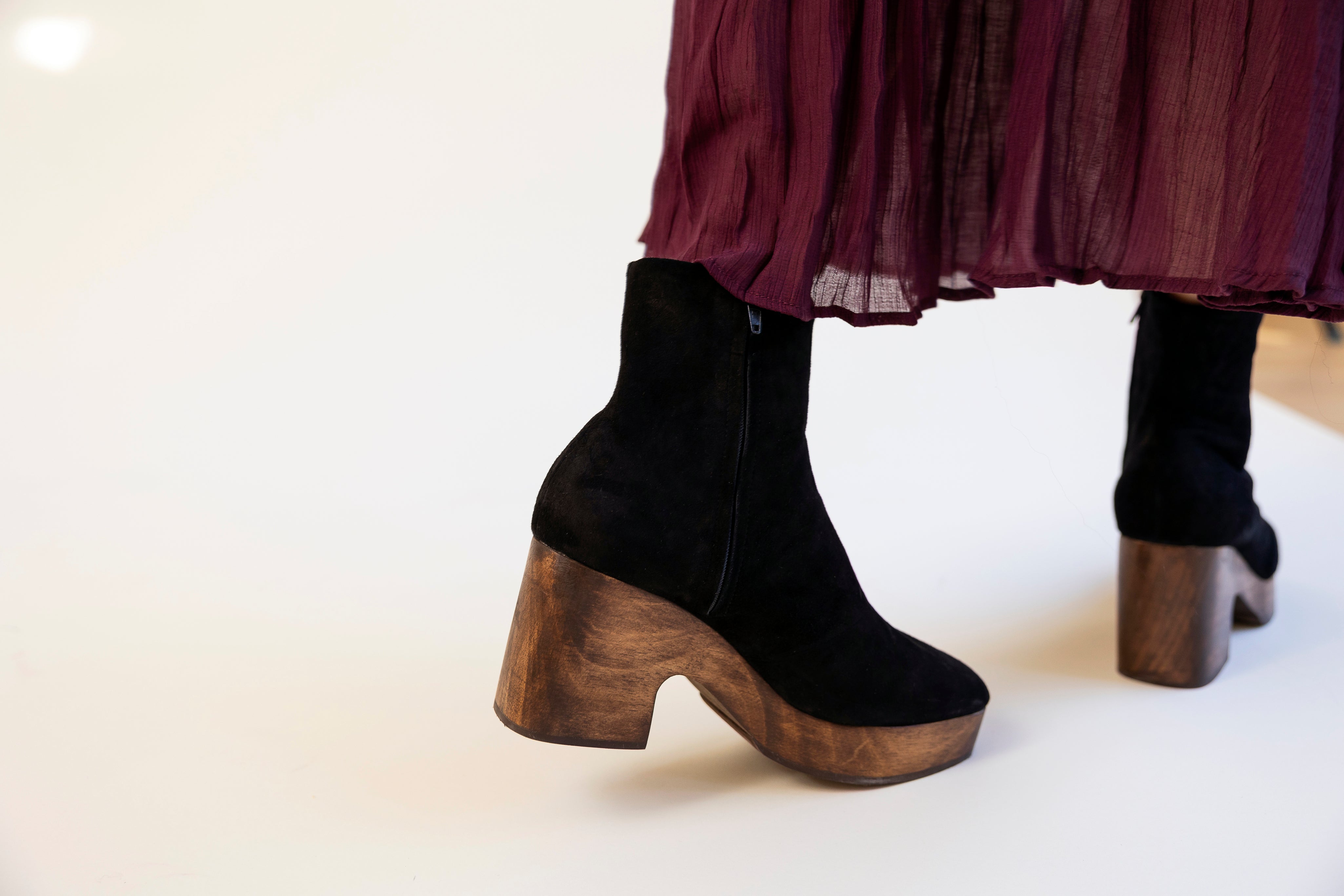 Black ankle boots with wooden soles worn by a person in a burgundy skirt on a white background