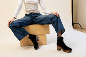 Person wearing blue jeans and black suede ankle boots sitting on a wooden box.
