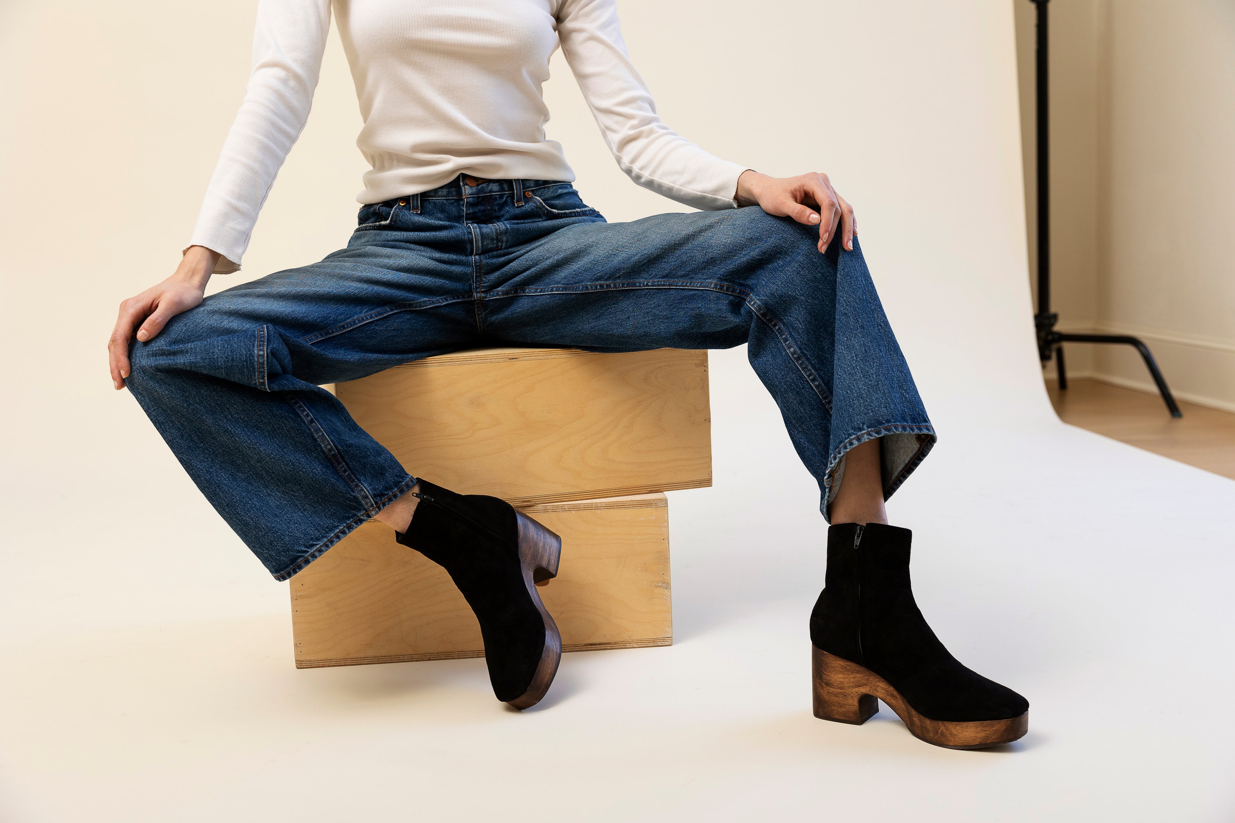 Person wearing blue jeans and black suede ankle boots sitting on a wooden box.