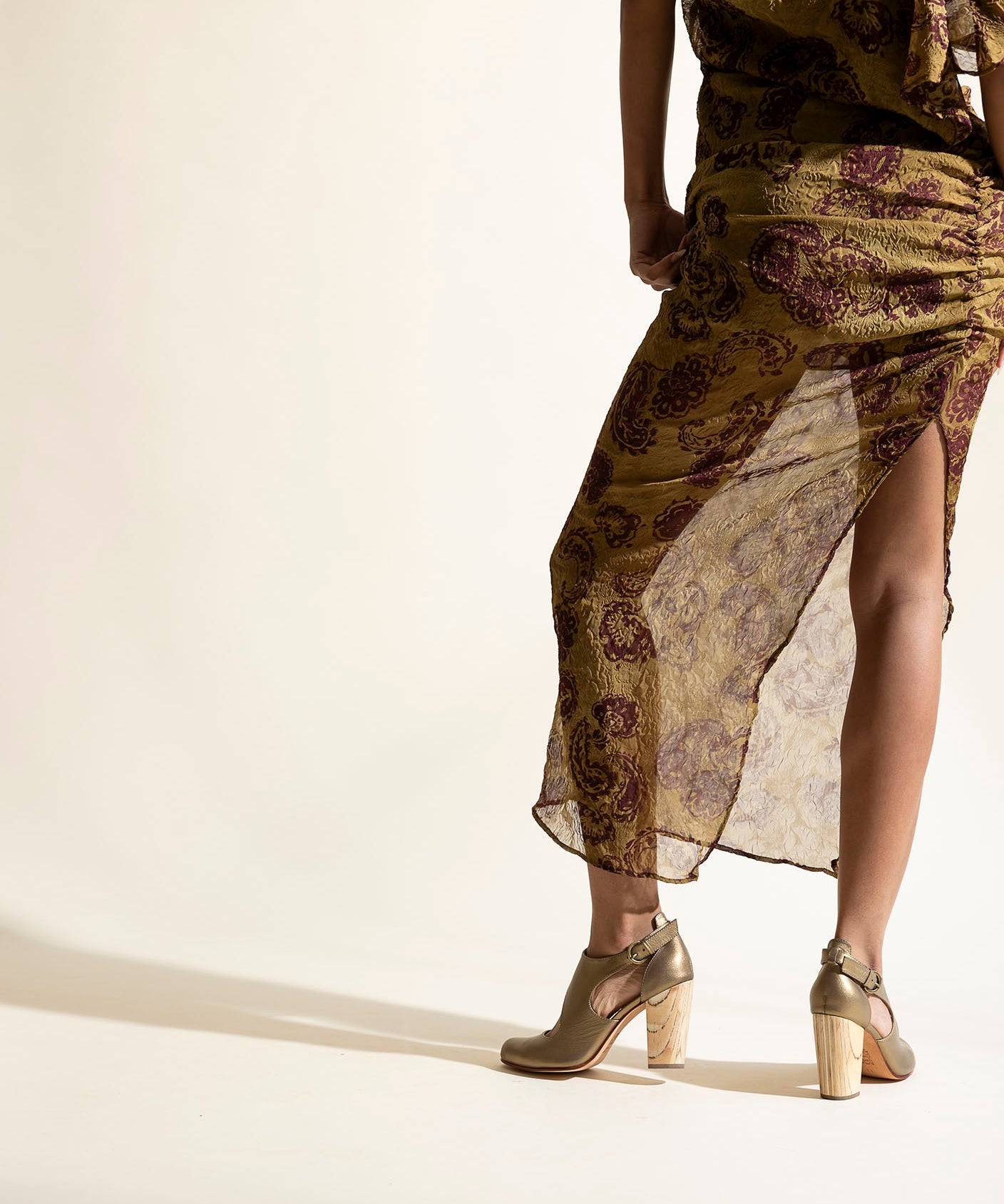 Woman wearing a long, patterned dress, gold heels, with sheer overlay on a beige background