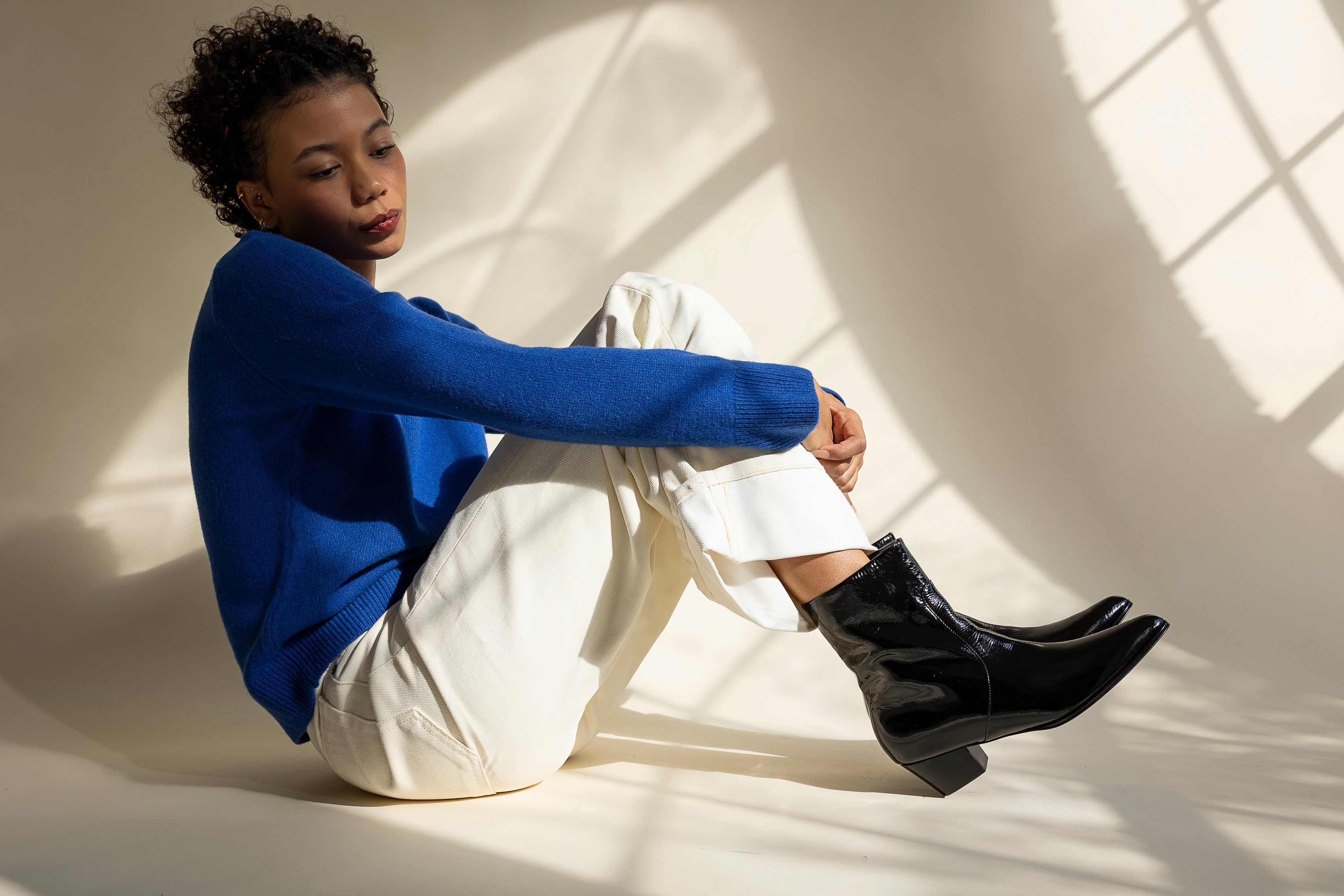 Person wearing a blue sweater and white pants sitting on a white floor with shadows cast by a window.