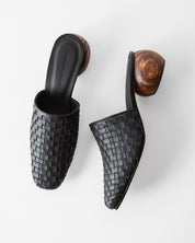 A pair of black woven leather mule sandals with chunky wooden block heels, shown from top-down angles on a light gray background.