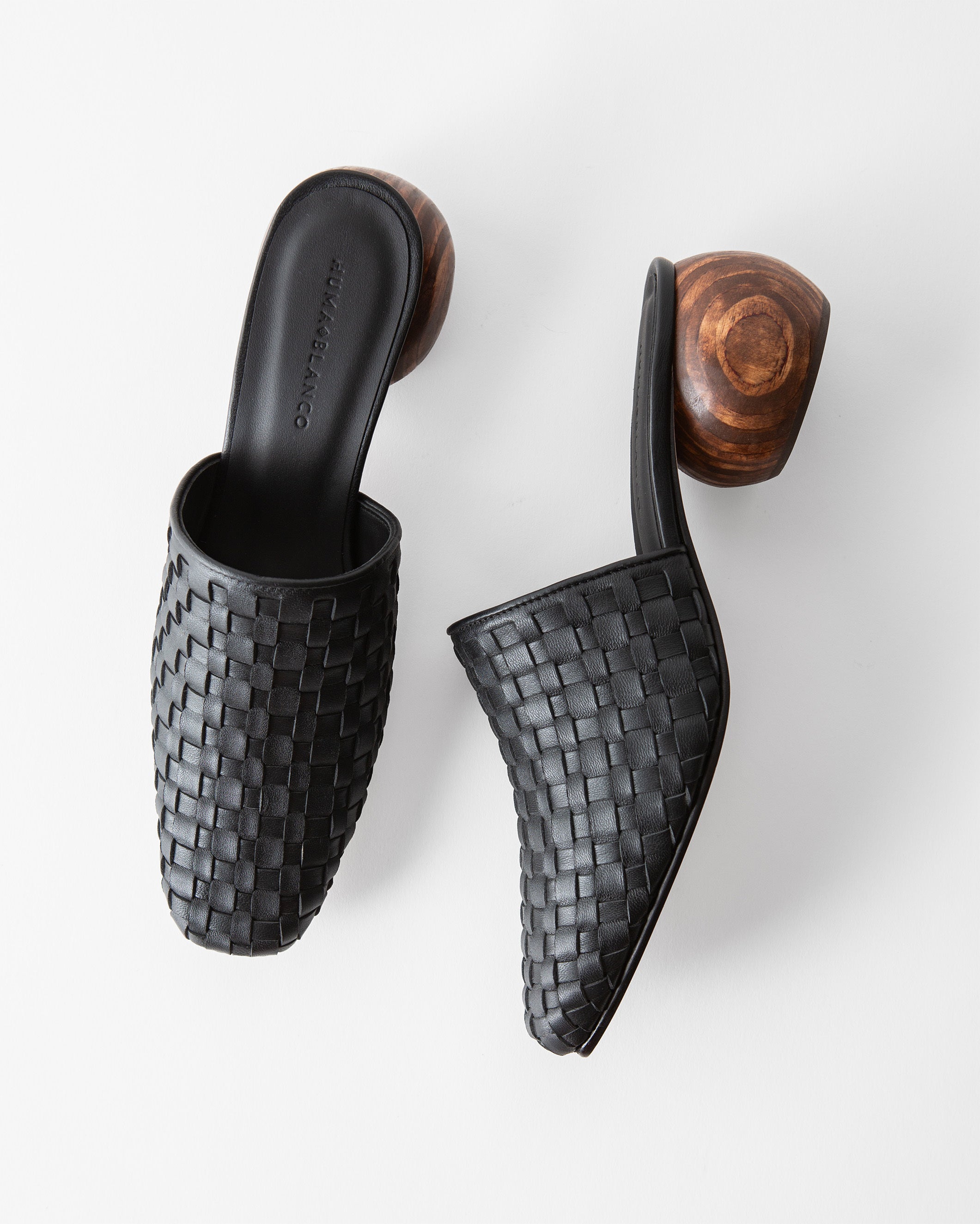 A pair of black woven leather mule sandals with chunky wooden block heels, shown from top-down angles on a light gray background.