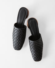 A pair of black woven leather mule sandals with square toes and brown stacked block heels, shown from a top-down angle on a light gray background.
