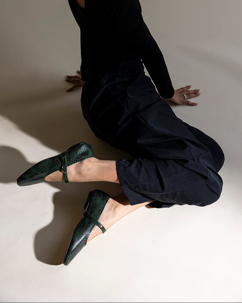 A model in a black long-sleeve top and black pants kneels on a warm beige studio floor, wearing dark green textured Mary Jane flats.