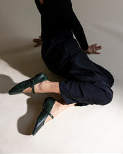 A model in a black long-sleeve top and black pants kneels on a warm beige studio floor, wearing dark green textured Mary Jane flats.