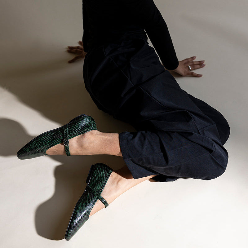 A model in a black long-sleeve top and black pants kneels on a warm beige studio floor, wearing dark green textured Mary Jane flats.