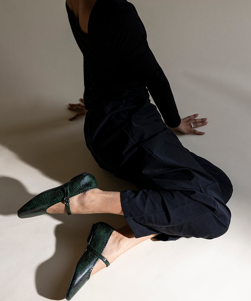 A model in a black long-sleeve top and black pants kneels on a warm beige studio floor, wearing dark green textured Mary Jane flats.