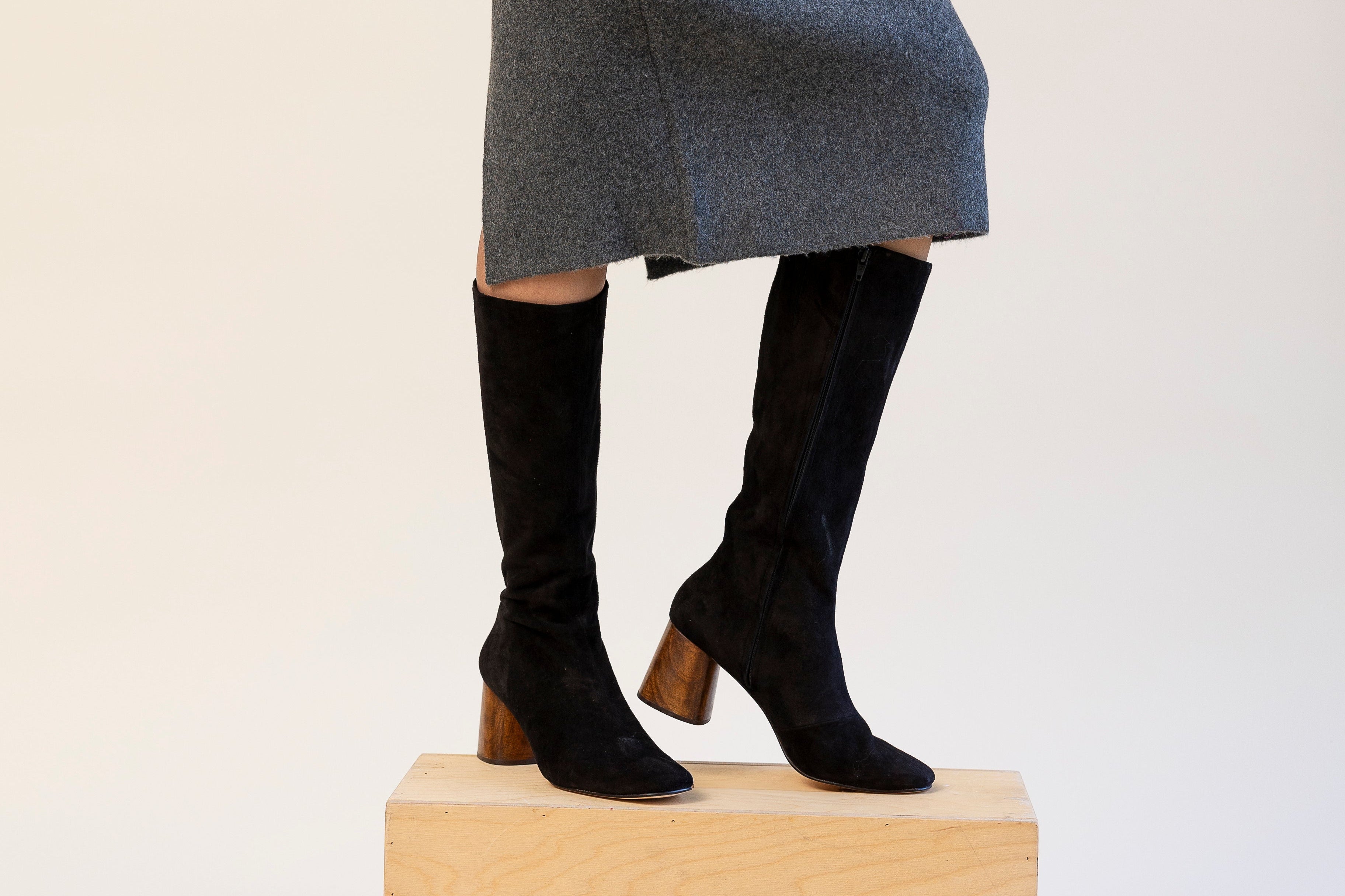 Person wearing a gray skirt and black knee-high boots on a beige block against a white background