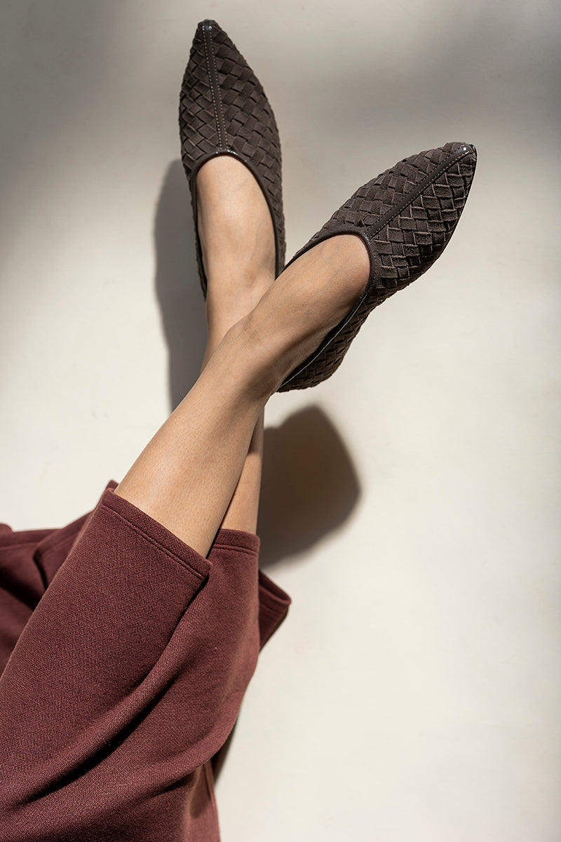 A close-up of a person's legs crossed, wearing dark brown woven pointed-toe flats and a matching dark brown wide-leg sweater, set against a warm off-white background with dramatic shadows.