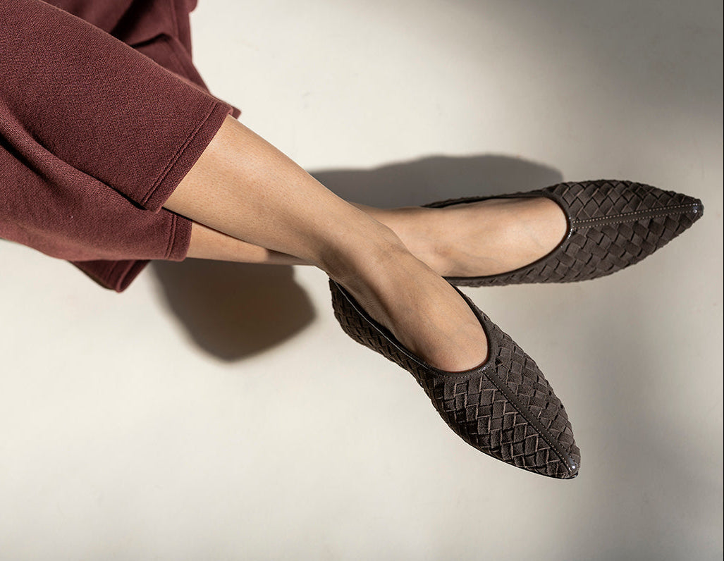 A close-up of a person's legs crossed, wearing dark brown woven pointed-toe flats and a matching dark brown wide-leg sweater, set against a warm off-white background with dramatic shadows.