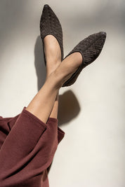 A close-up of a person's legs crossed, wearing dark brown woven pointed-toe flats and a matching dark brown wide-leg sweater, set against a warm off-white background with dramatic shadows.