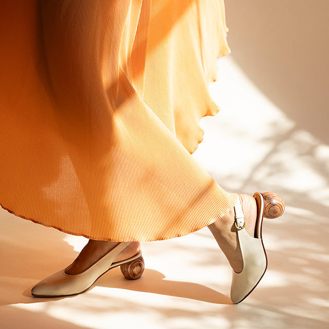 A close-up lifestyle shot of a person wearing a flowing orange dress and white slingback kitten heels with gold accents, walking on a sunlit beige floor with leaf shadows.